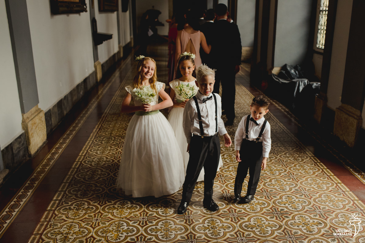 pajens usando camisa branca, gravata borboleta e suspensório preto e daminhas segurando buque verde e usando vestido branco posam para retrato 