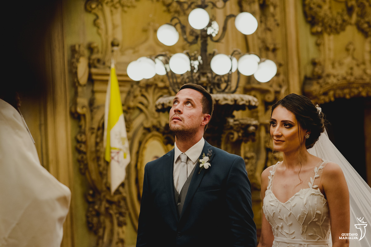 noivo usando terno azul e gravata cinza olha para cima enquanto noiva observa sermão do padre