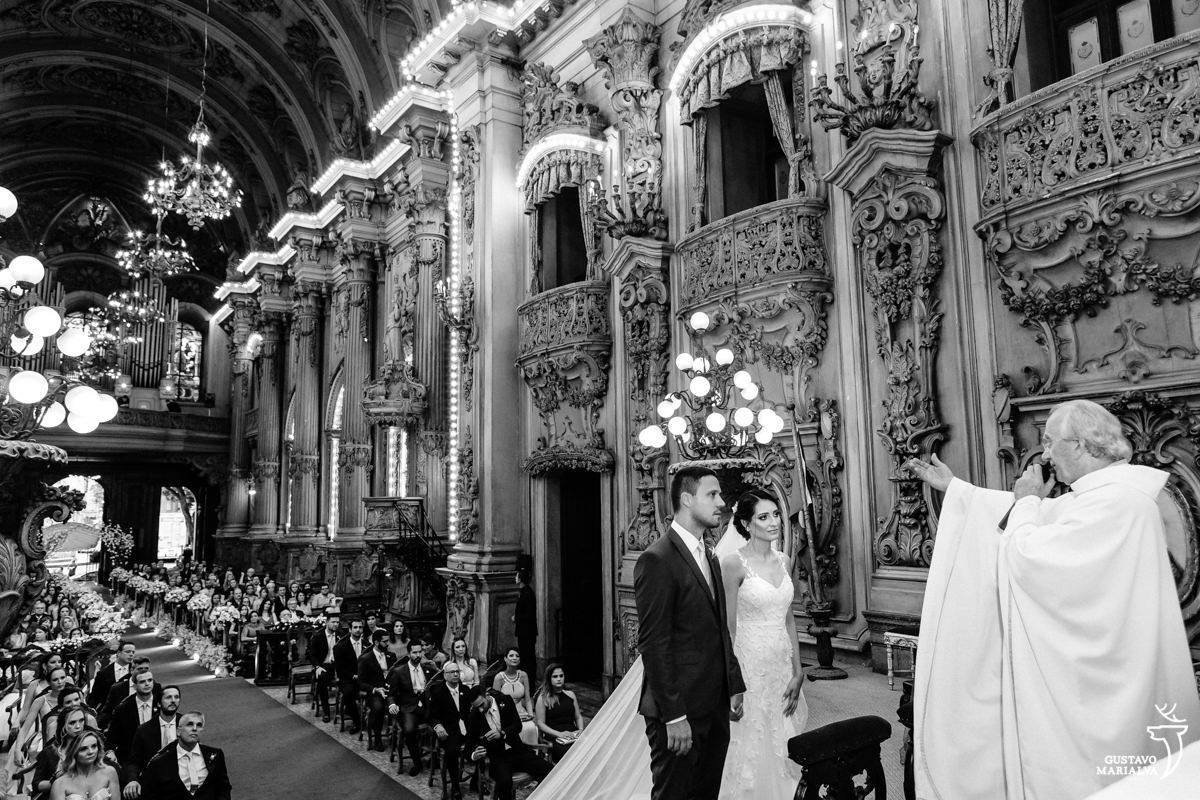padre abençoa o casal de noivos que observa a cerimônia com a igreja ao fundo