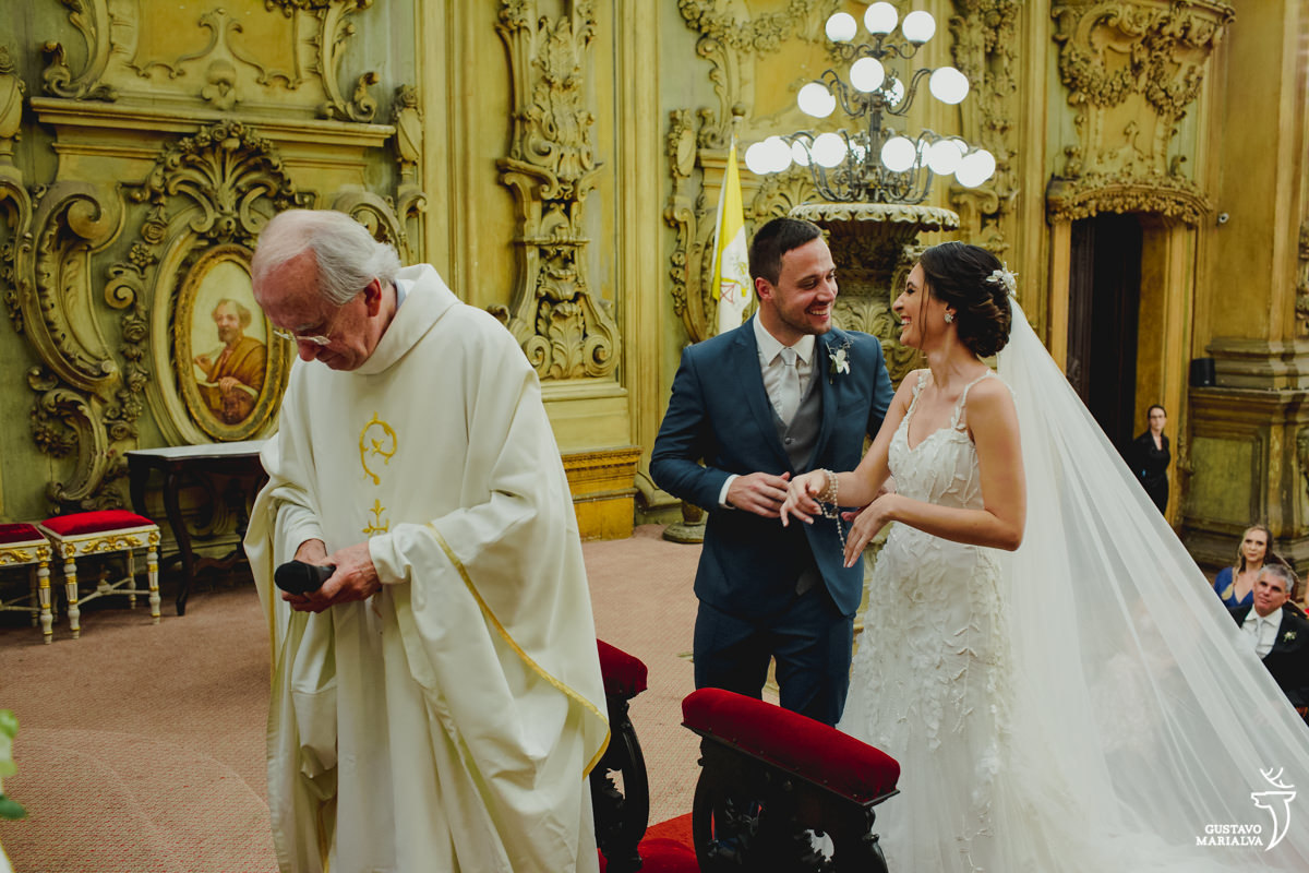 padre tenta desligar o celular enquanto noivos de mãos dadas sorriem