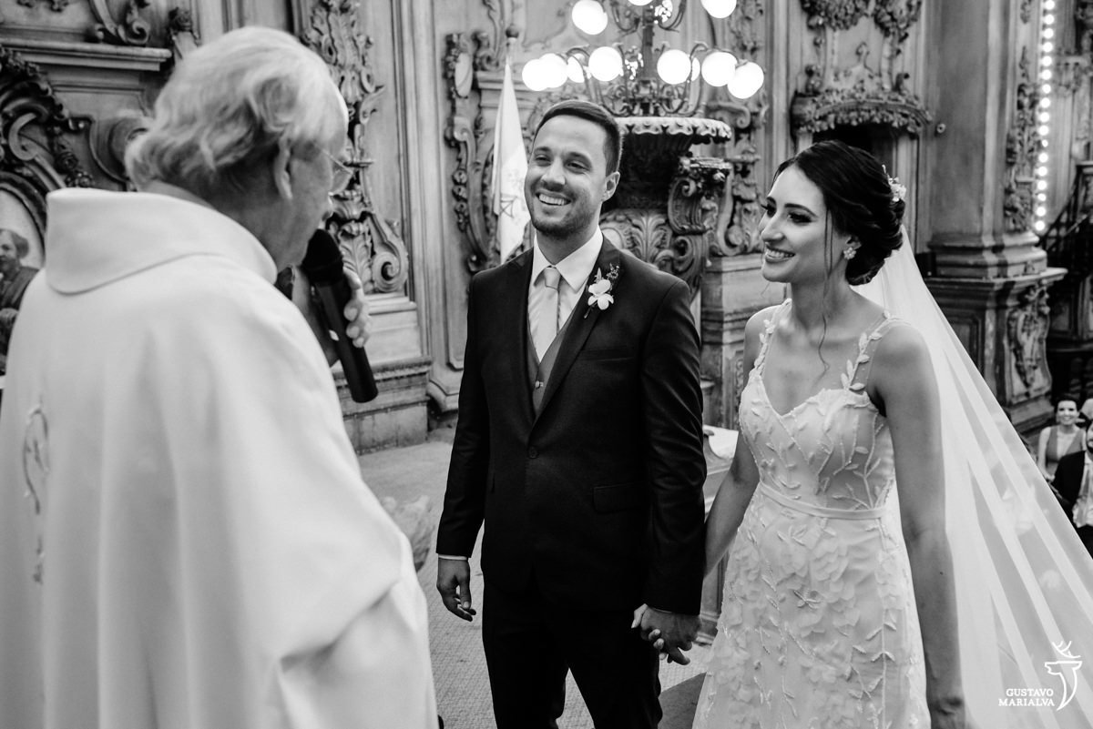 noivos de mãos dadas sorriem enquanto escutam o padre falar durante a cerimônia de casamento