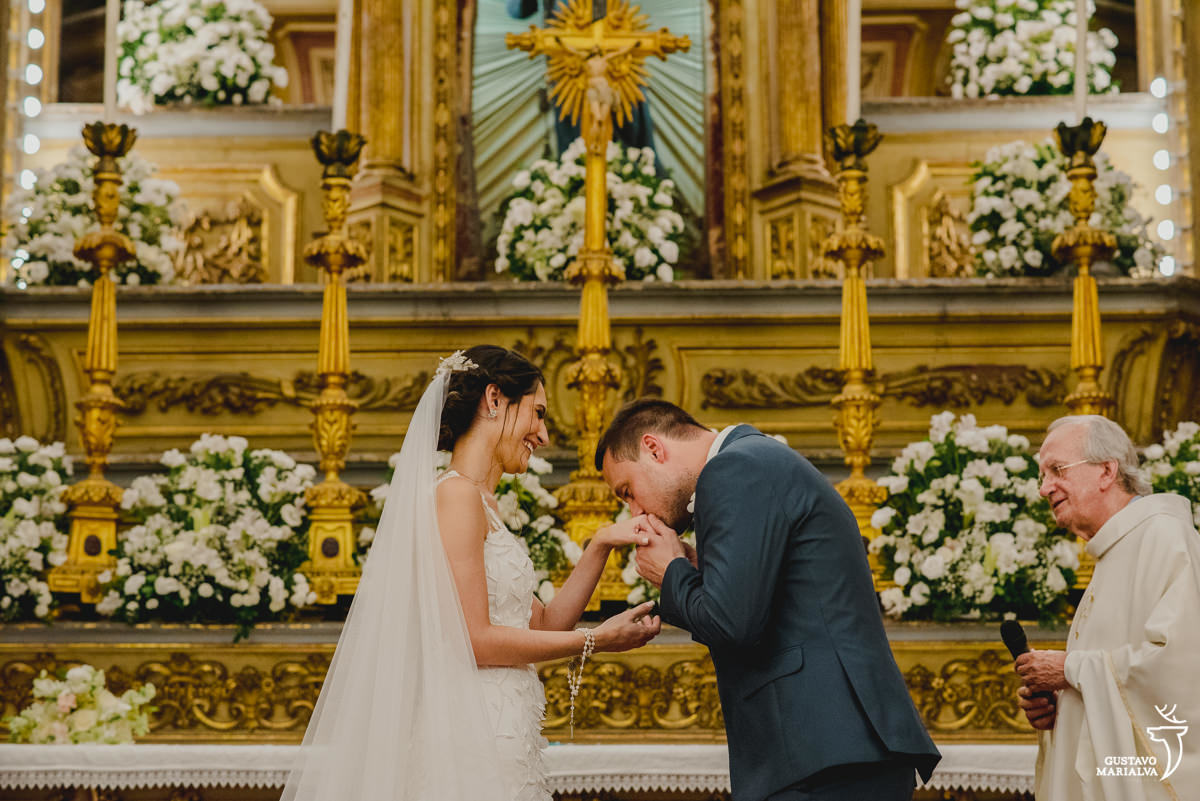 noivo beija a mão da noiva enquanto é observado pelo padre