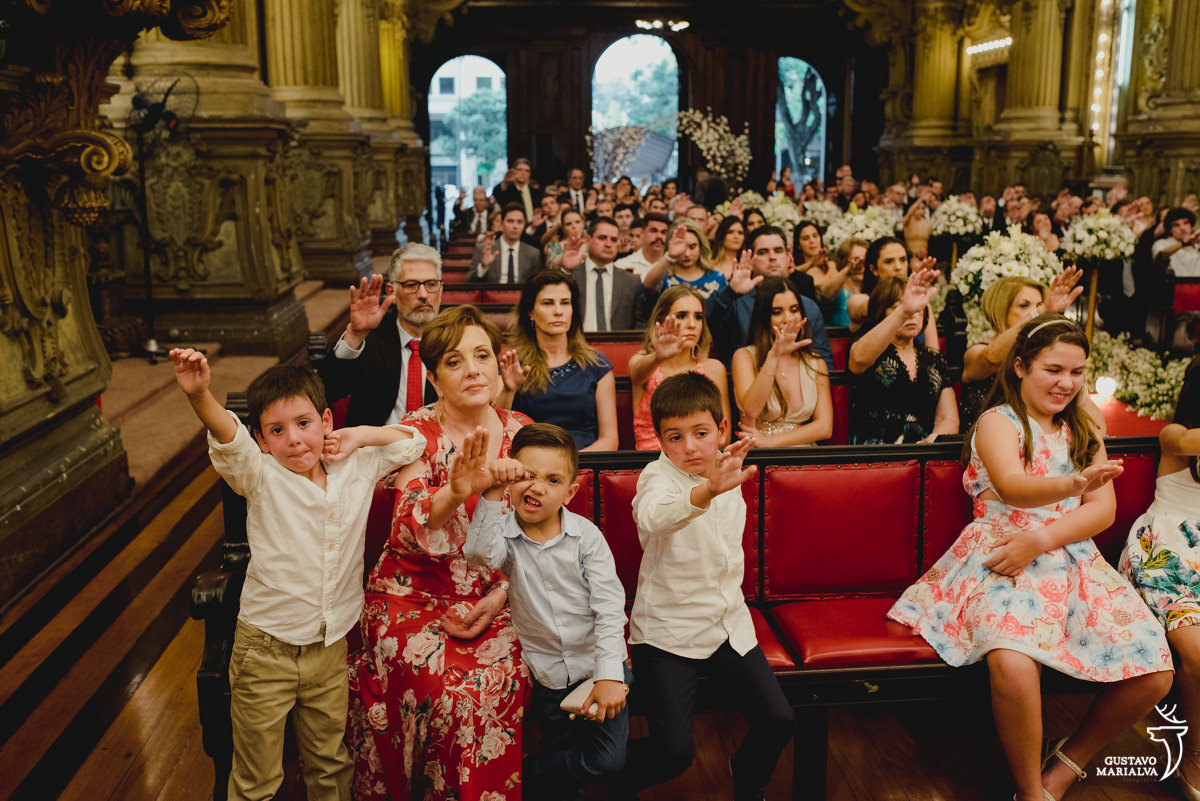 menino usando camisa azul faz careta enquanto menino usando camisa branca estica o braço e são observados por mulher que usa vestido florido