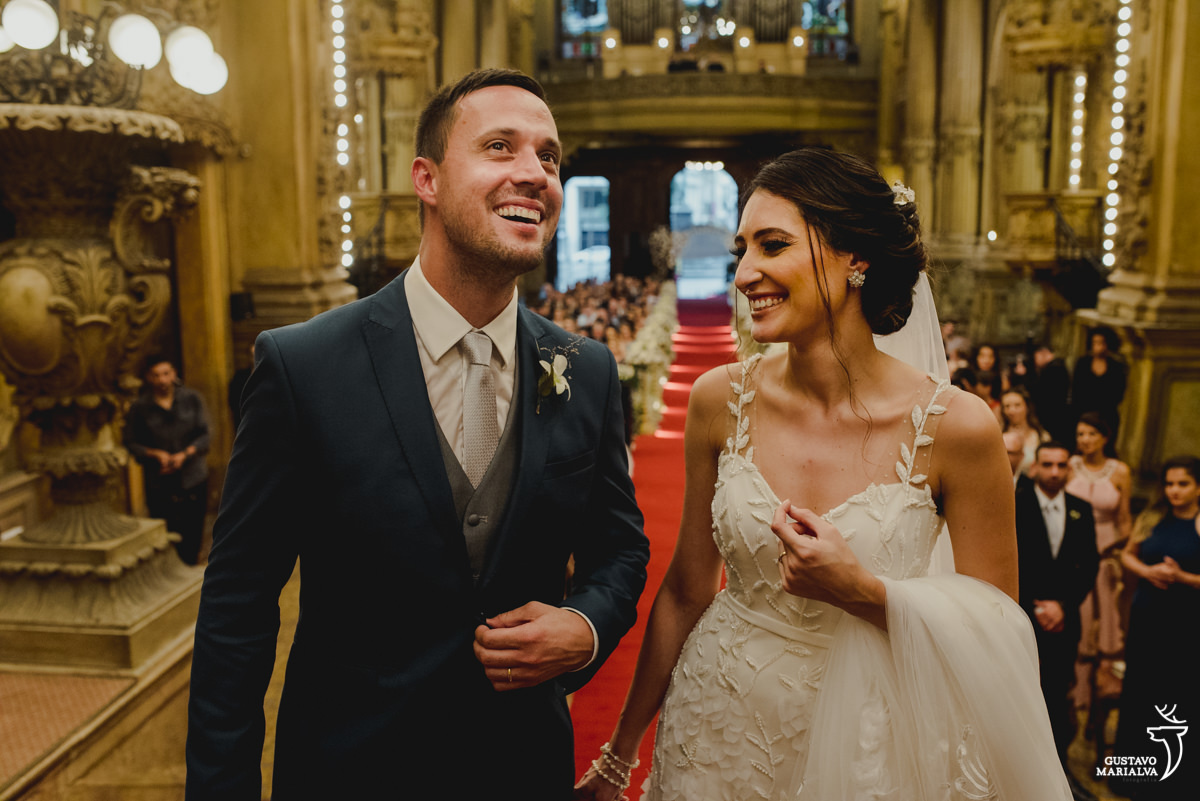 noiva segurando o véu sorri ao olhar para o noivo que olha para o teto