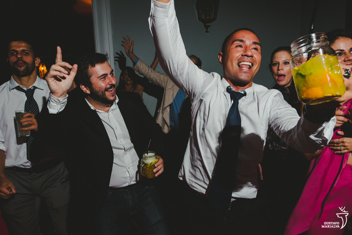 homem usando camisa branca e gravata azul dança segurando bebida amarela enquanto é acompanhado por amigo que usa camisa branca e blazer preto