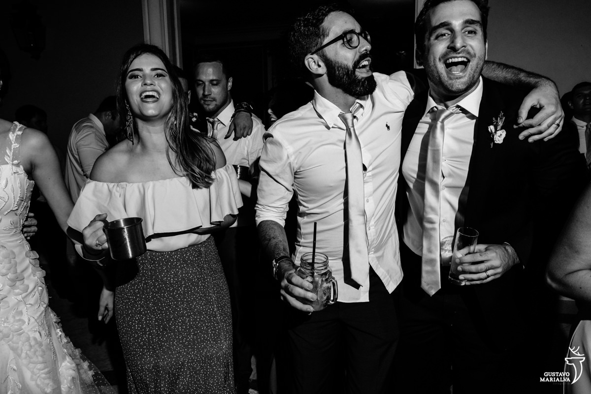 convidado usando camisa branca e gravata prata dança com amigo enquanto é acompanhado por mulher que usa vestido rosa