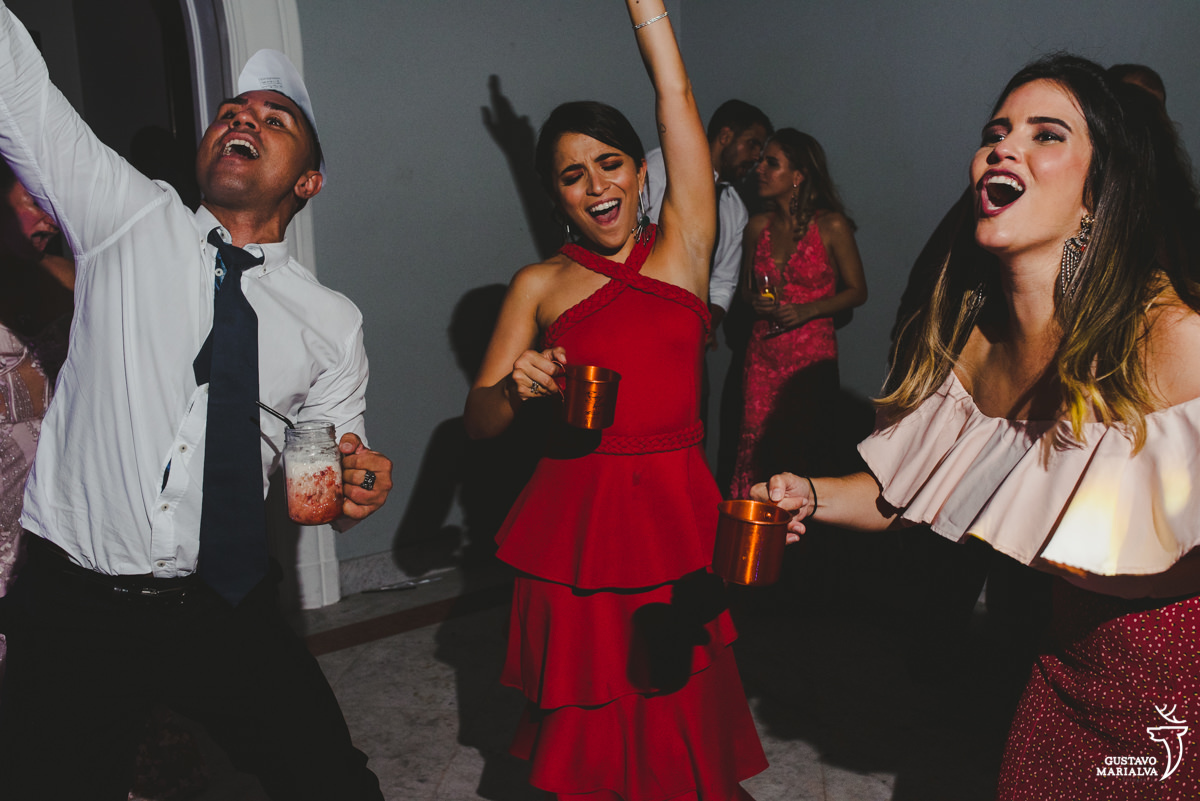 convidada usando vestido vermelho e segurando caneca dança com os braços para cima enquanto homem usando camisa branca e gravata preta dança animado 