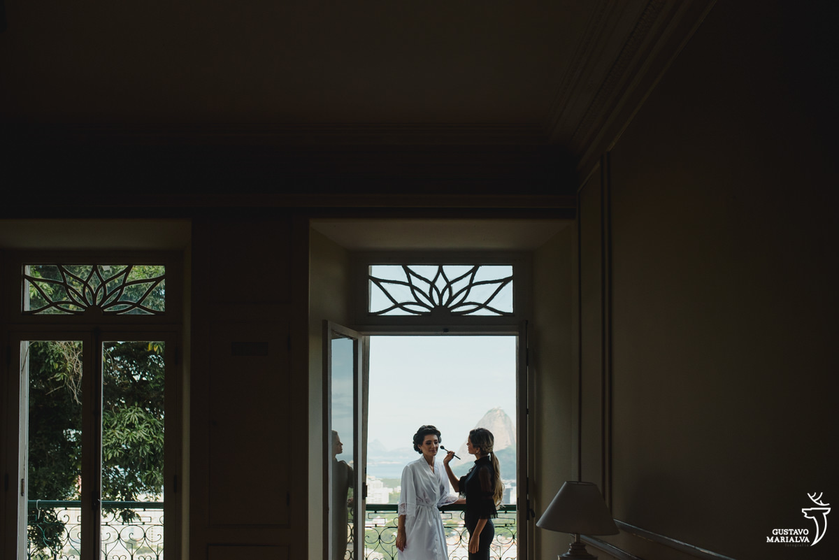 maquiadora passando batom na noiva na varanda da casa com o pão de açúcar ao fundo