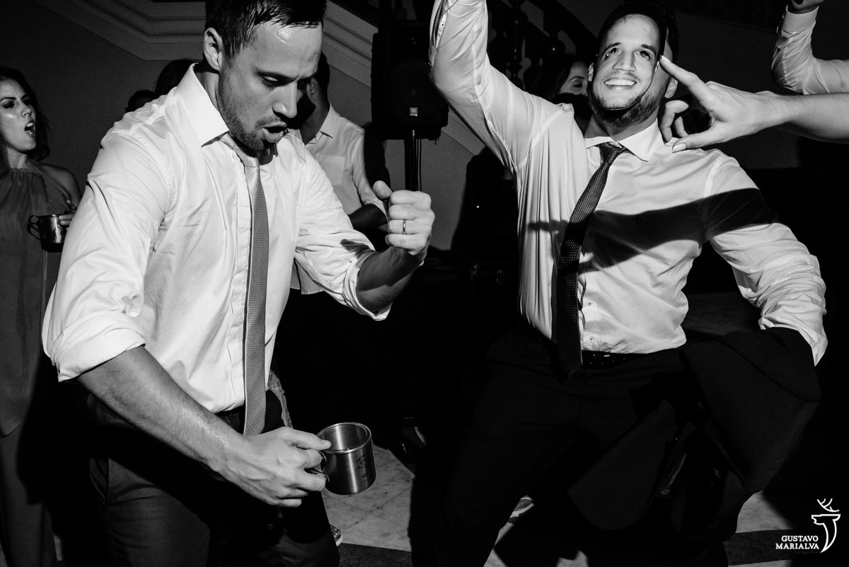 noivo dança segurando caneca enquanto é acompanhado por amigo que usa camisa branca, gravata e máscara
