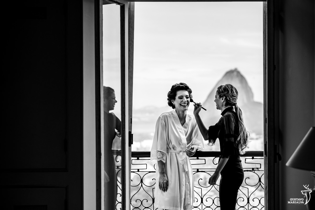 noiva usando robe branco sorri enquanto maquiadora passa blush com a imagem do pão de açúcar ao fundo