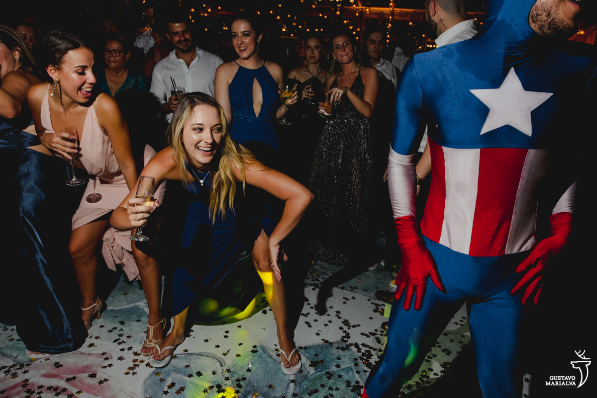 mulher loira de vestido azul dança rindo com a pose do capitão américa