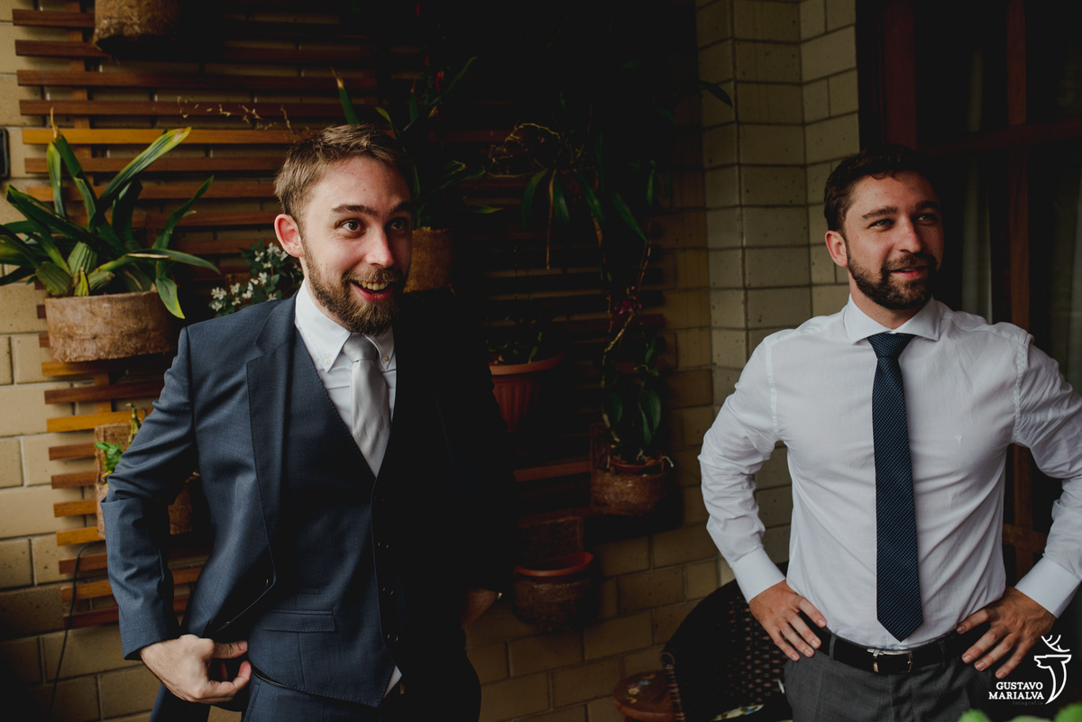 noivo sorri enquanto arruma o paletó cinza escuro e é observado pelo irmão