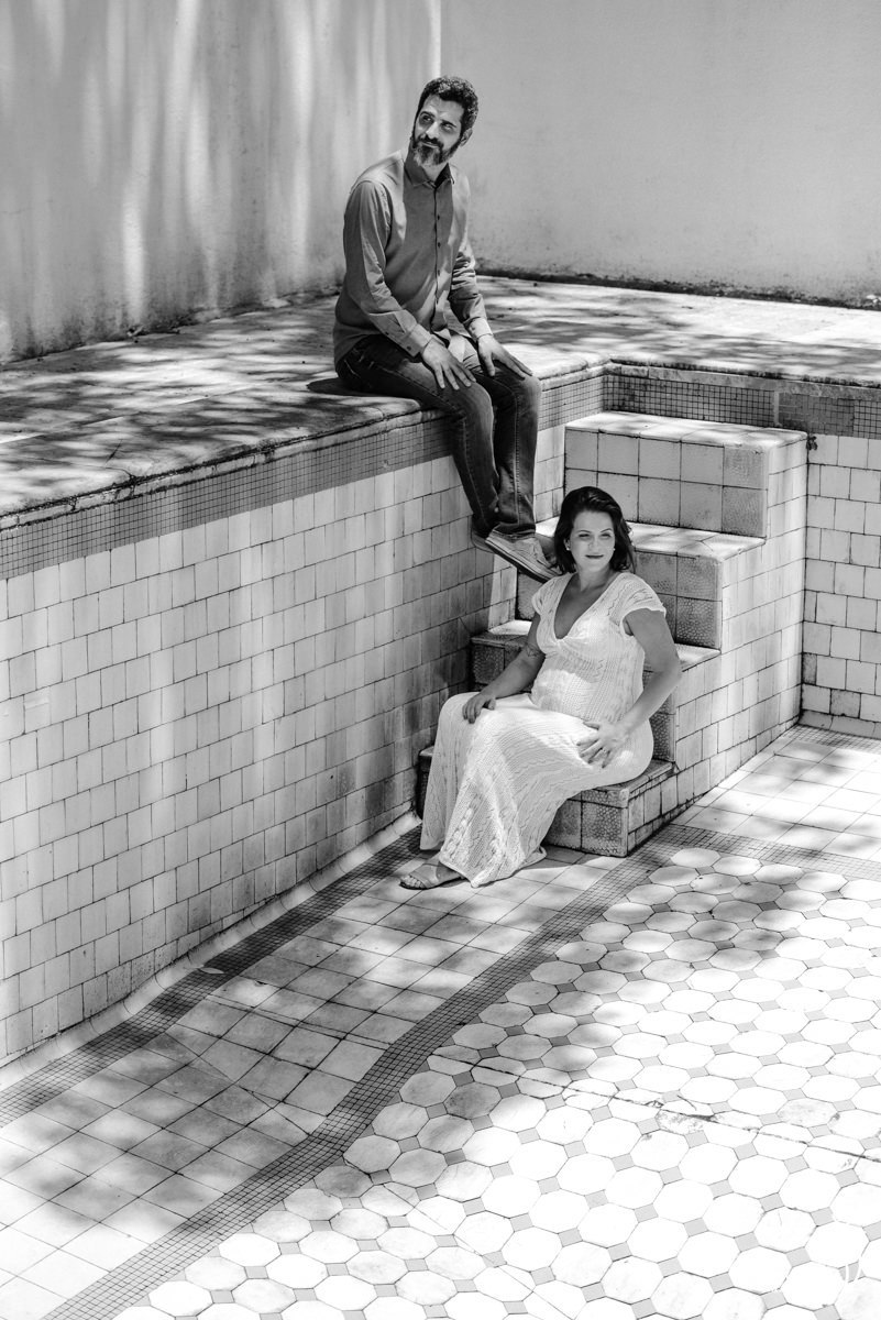 homem sentado na borda da piscina observa a esposa grávida sentada na escada da piscina vazia