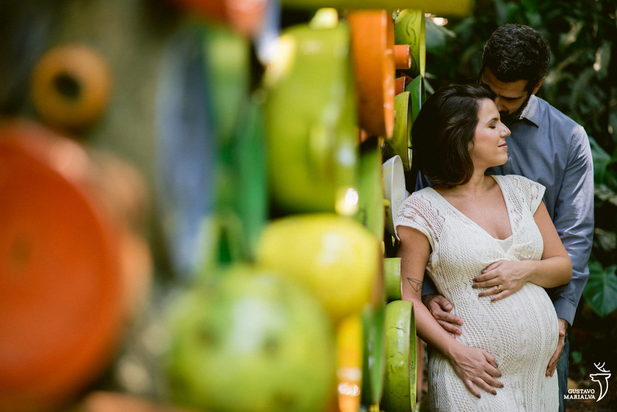 grávida usando vestido branco é abraçada pelo marido que usa camisa azul com fundo verde