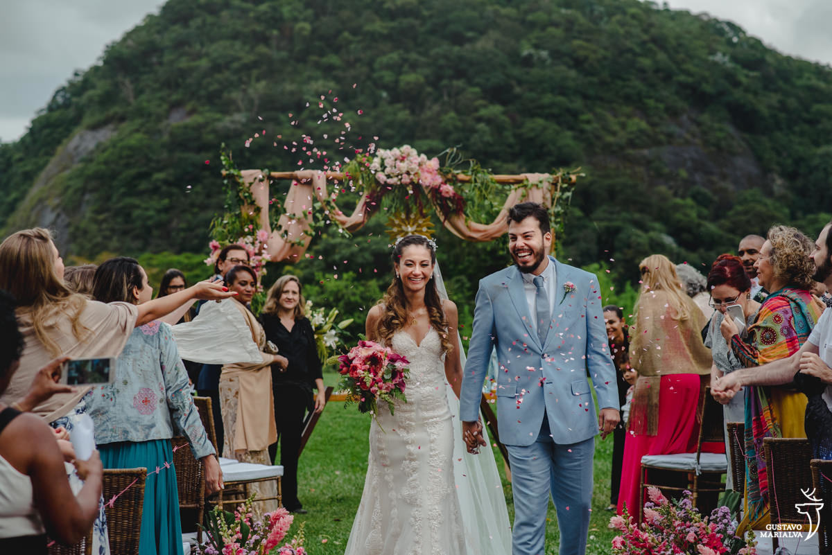 noivos saem de mãos dadas enquanto convidados jogam pétalas de rosa para cima