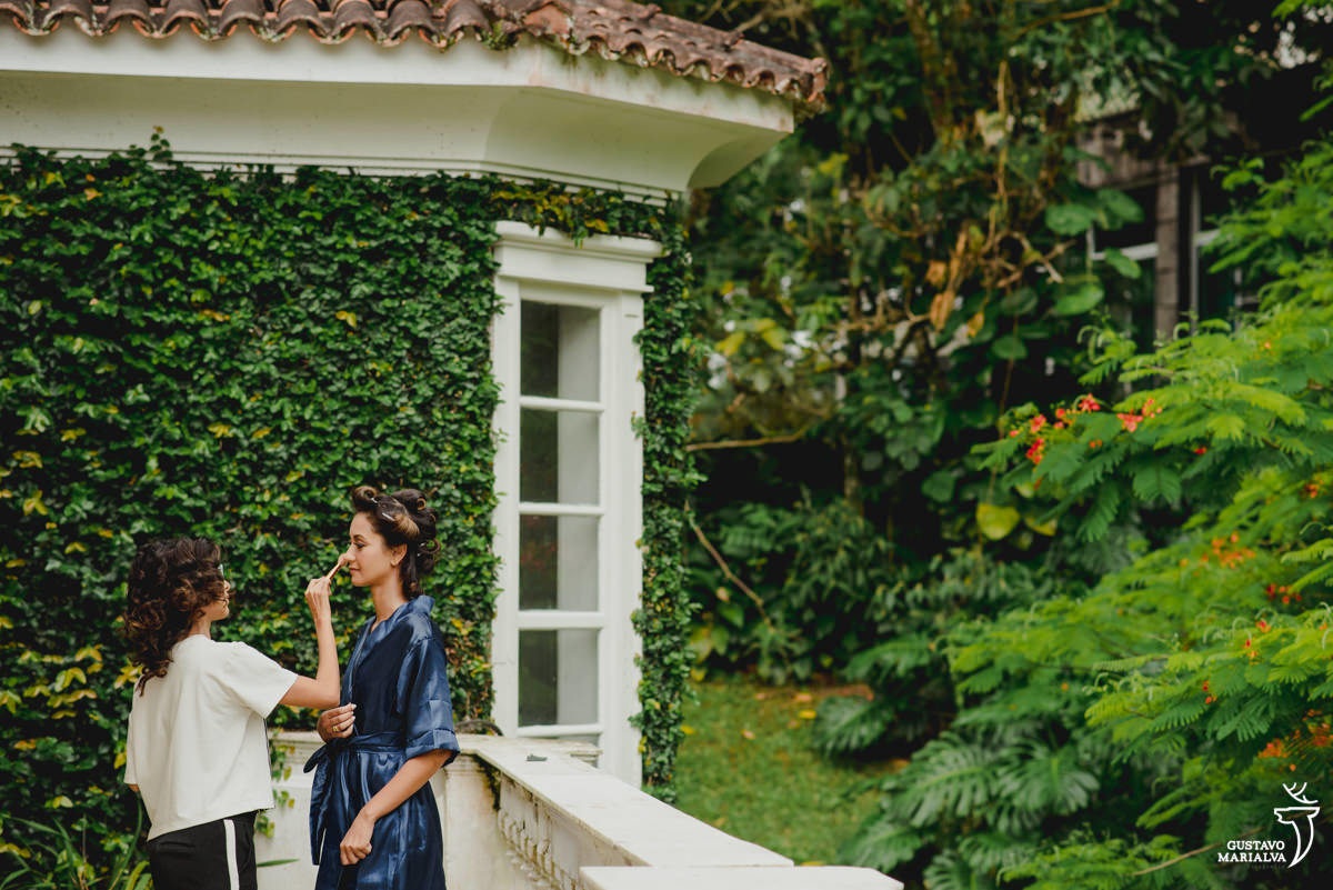 noiva passando blush usando robê azul marinho na varanda da casa em frente a parede de plantas verdes