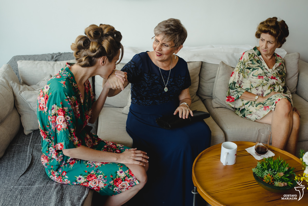 mulher usando robe florido beija a mão de senhora que usa vestido azul e é observada pela mãe que usa rove florido