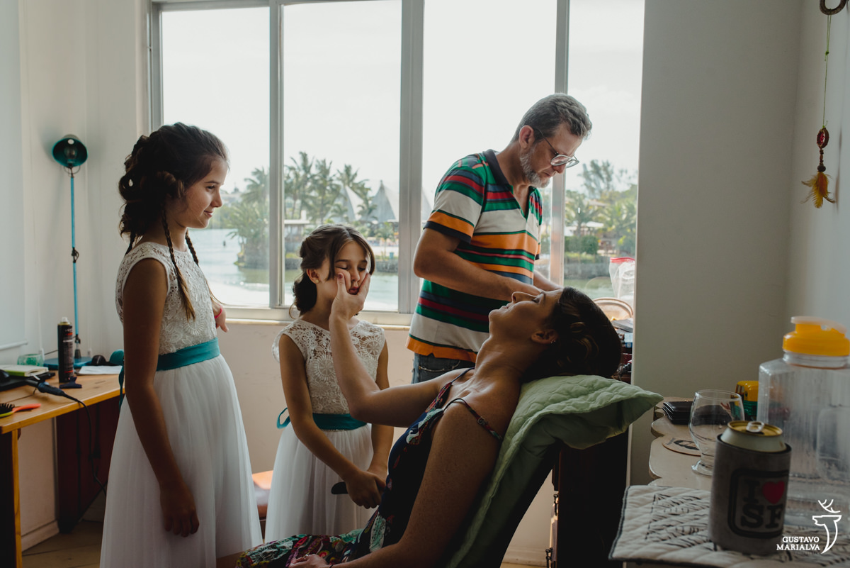 mulher sendo maquiada aperta a bochecha da filha que usa vestido branco com faixa azul