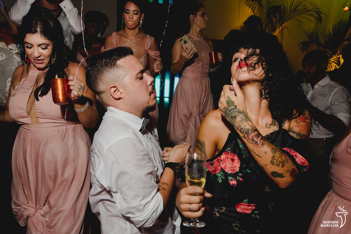 mulher morena de batom vermelho e braços tatuados dança com homem que usa camisa branca e gravata borboleta