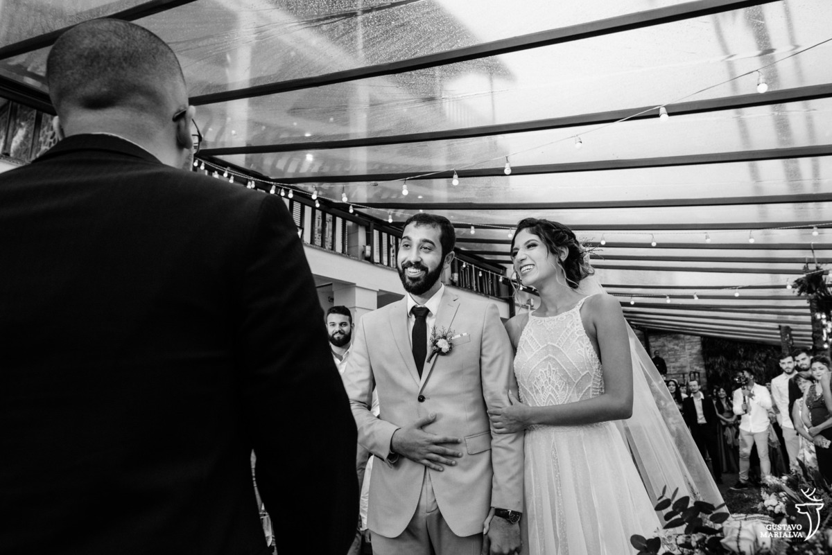 noivos sorriem e observam amigo que usa terno preto celebrar a cerimônia do casamento