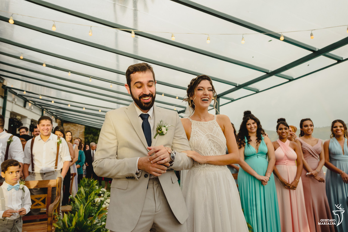 noivos de mãos dadas sorriem enquanto são observados por madrinhas que usam vestido rosa e verde