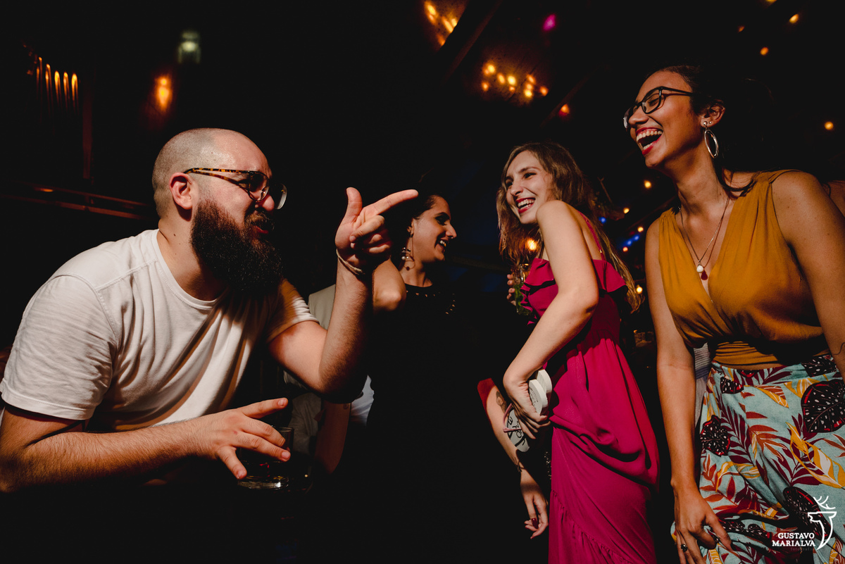 homem careca e usando camisa branca dança e aponta para amiga que sorri