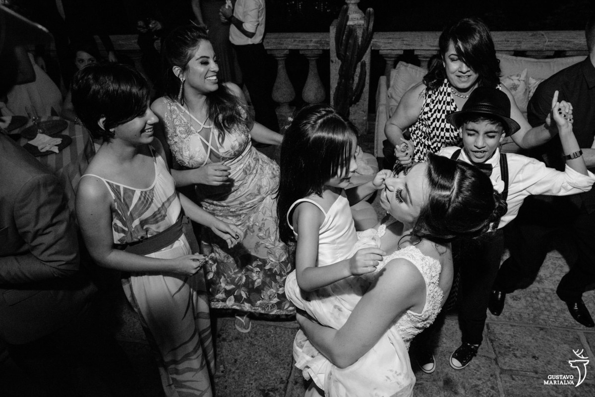 noiva carrega daminha no colo enquanto menino de camisa branca e chapéu preto dança ao fundo acompanhado de madrinha que usa vestido rosa