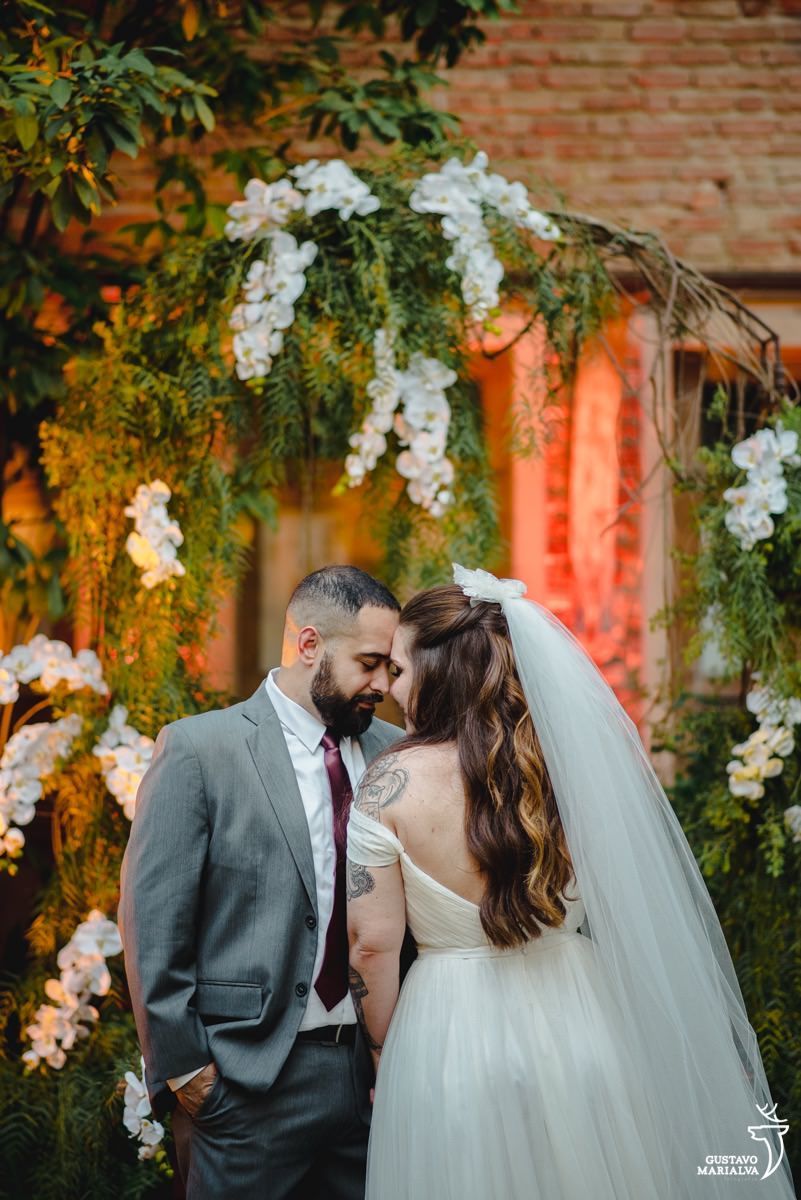 noivo usando terno cinza, camisa branca e gravata vinho dá as mãos para a noiva com flores e parede de tijolos ao fundo