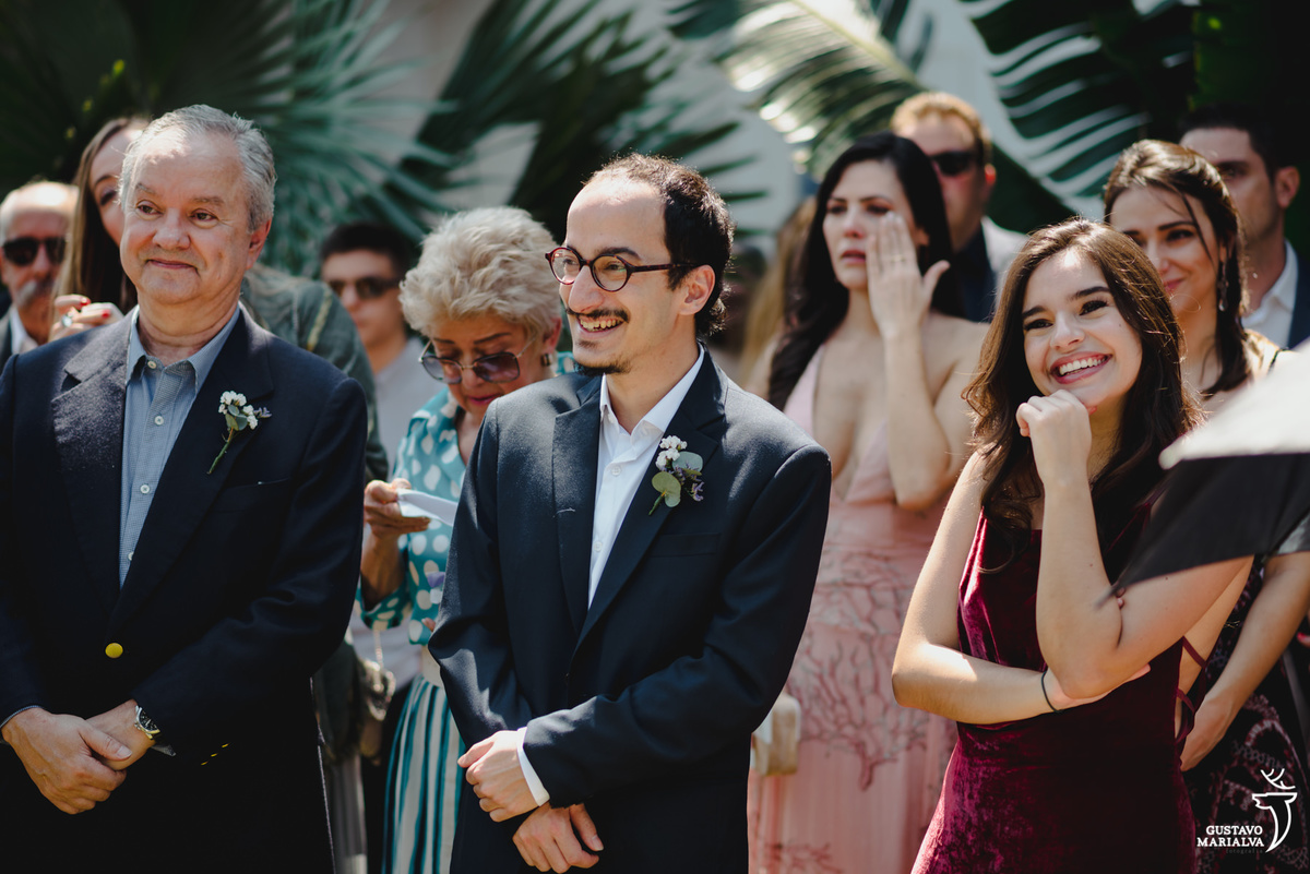 irmã do noivo usando vestido vinho sorri emocionada enquanto amigo usando terno cinza sorri