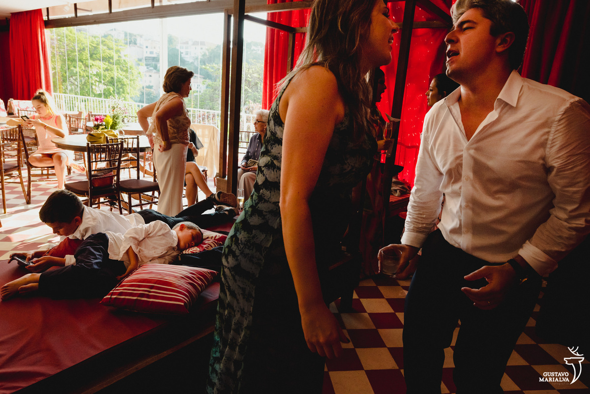 menino dorme em cama vermelha enquanto outro usando camisa branca joga video game e casal dança em frente a eles