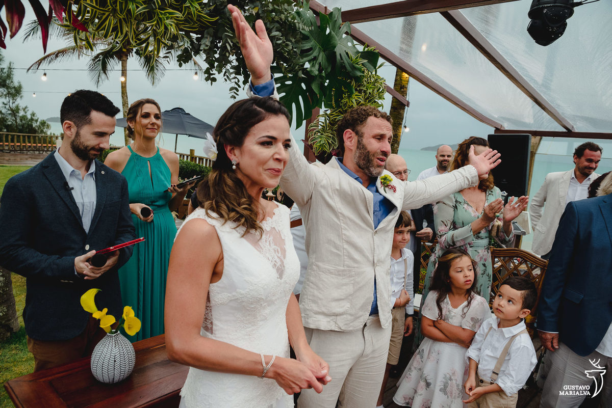 noivo abre os braços usando terno caqui e camisa azul enquanto noiva chora