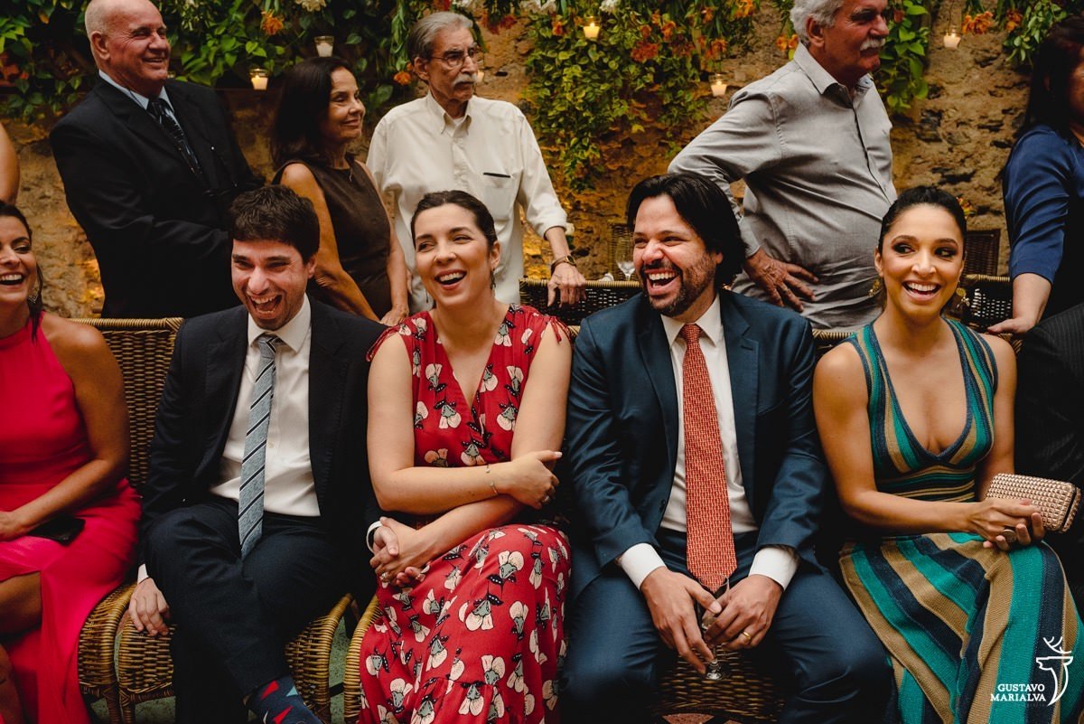 padrinhos usando terno azul e gravata cinda e laranja sorriem enquanto madrinha usando vestido azul, verde e preto se emociona