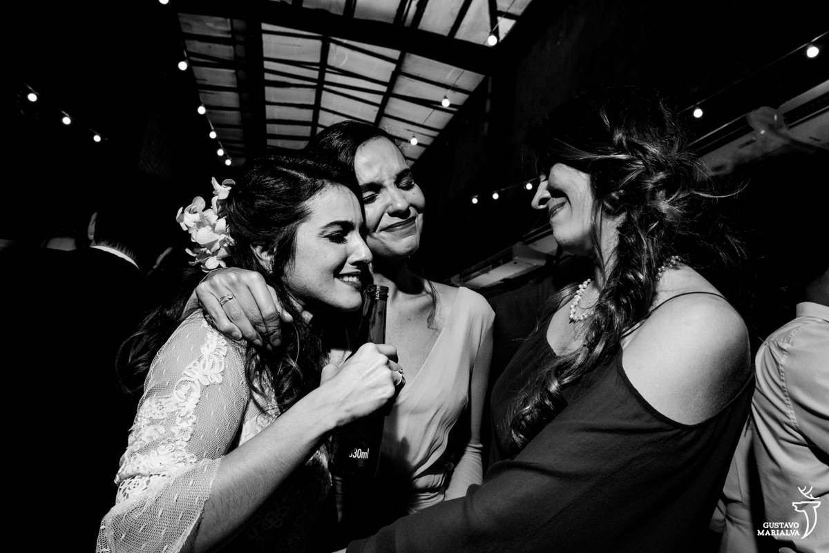 noiva segurando garrafa de cerveja chora ao abraçar madrinhas que usam vestido laranja e vinho