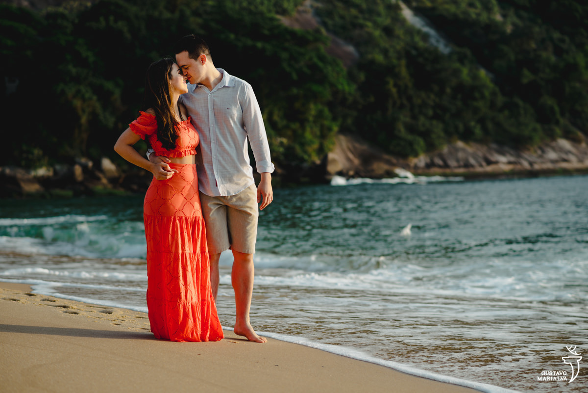 noiva de vestido laranja caminha abraçada ao namorado de camisa azul na praia vermelha