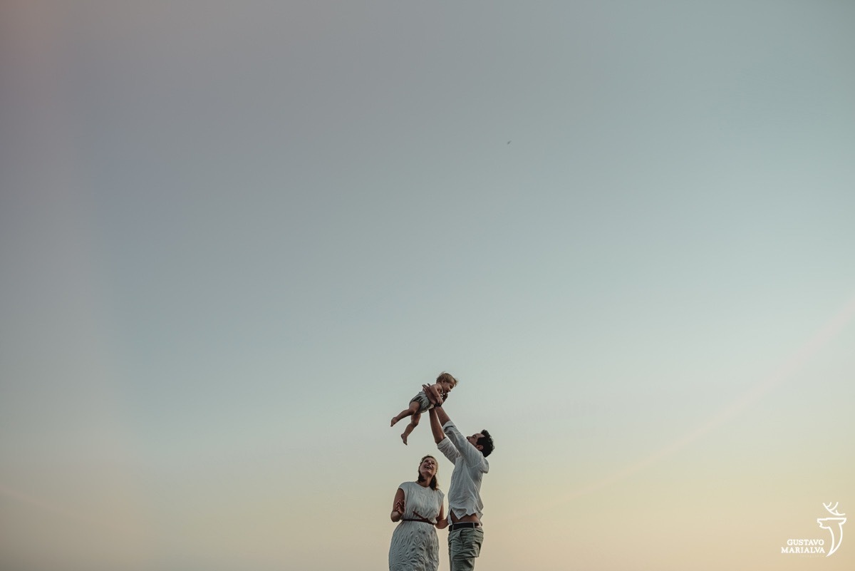 pai usando camisa branca e calça bege joga o filho para cima enquanto mãe sorri com por do sol ao fundo