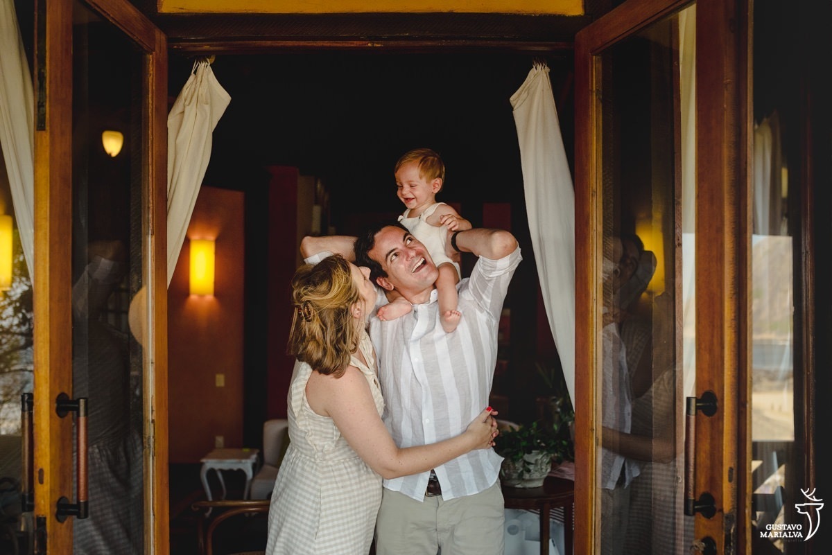 criança de macacão bege sorri sentada na corcunda do pai que usa calça bege e camisa branca e bege listrada enquanto mãe que usa vestido bege abraça