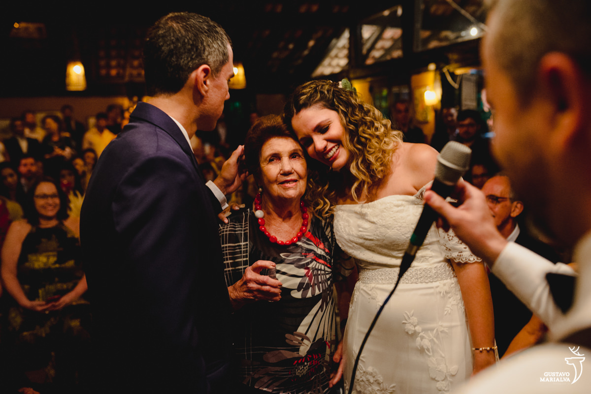 noiva abraça a avó emocionada que usa vestido estampado cinza e vermelho enquanto noivo que usa terno azul faz carinho 