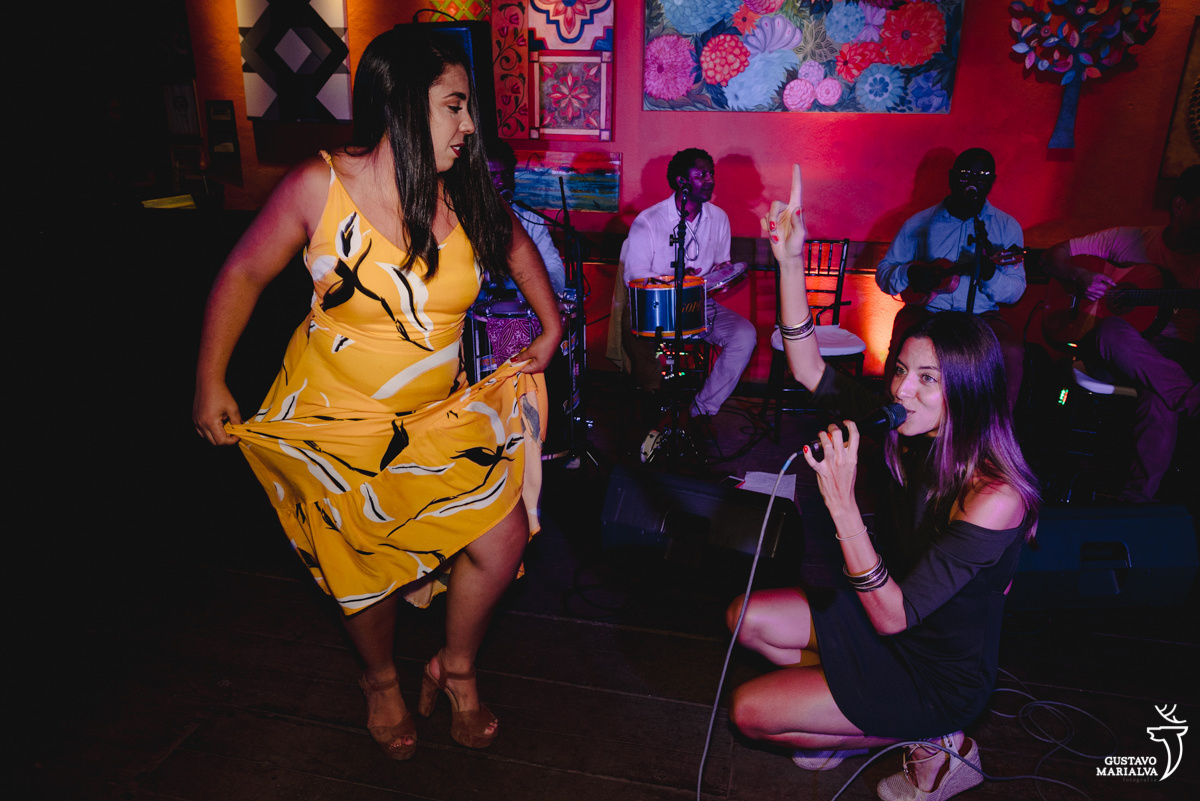mulher usando vestido amarelo estampado samba enquanto cantora de vestido verde canta agachada