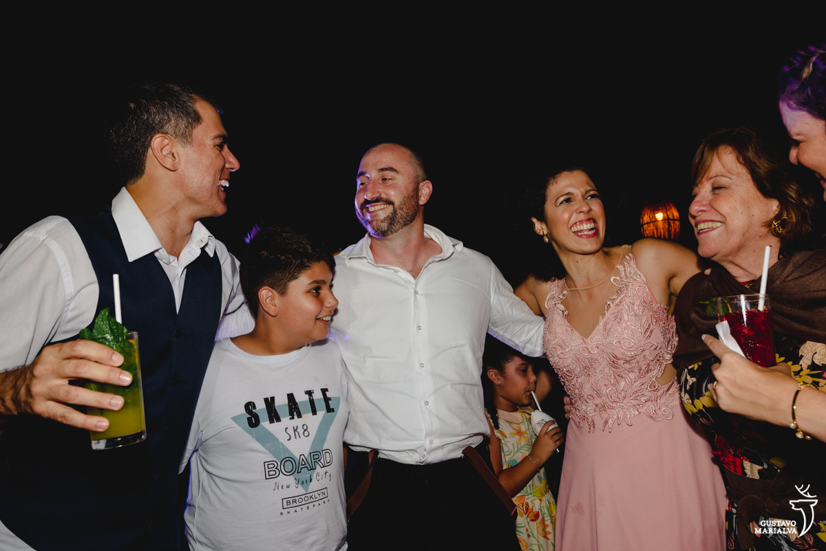 noivo segurando copo de bebida dança abraçado com menino de camisa branca e convidados que usam camisa branca e vestido rosa