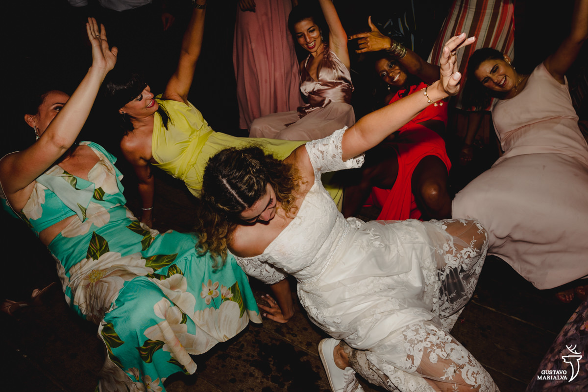 noiva dança agachada no chão acompanhada de amigas de vestido amarelo, estampado e vermelho