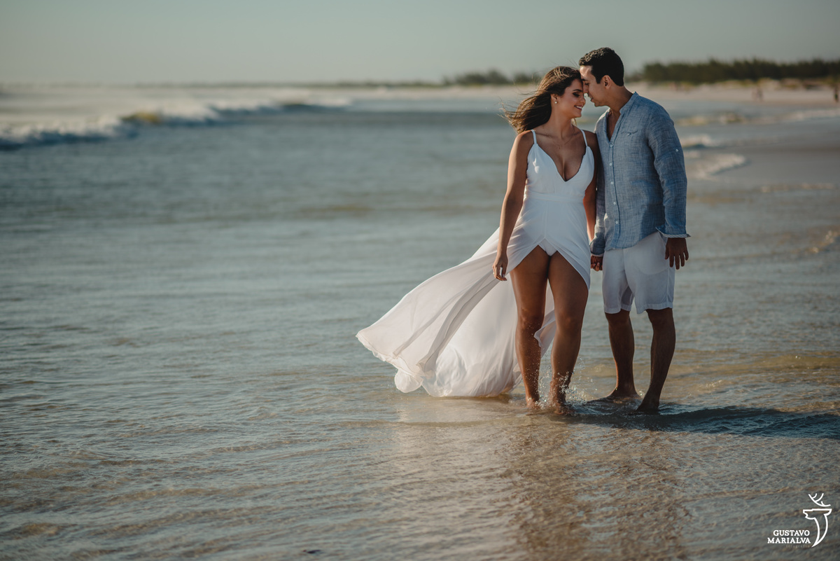 casal caminha de mãos dadas na praia grande em arraial do cabo e vento voa a calda do vestido da noiva 