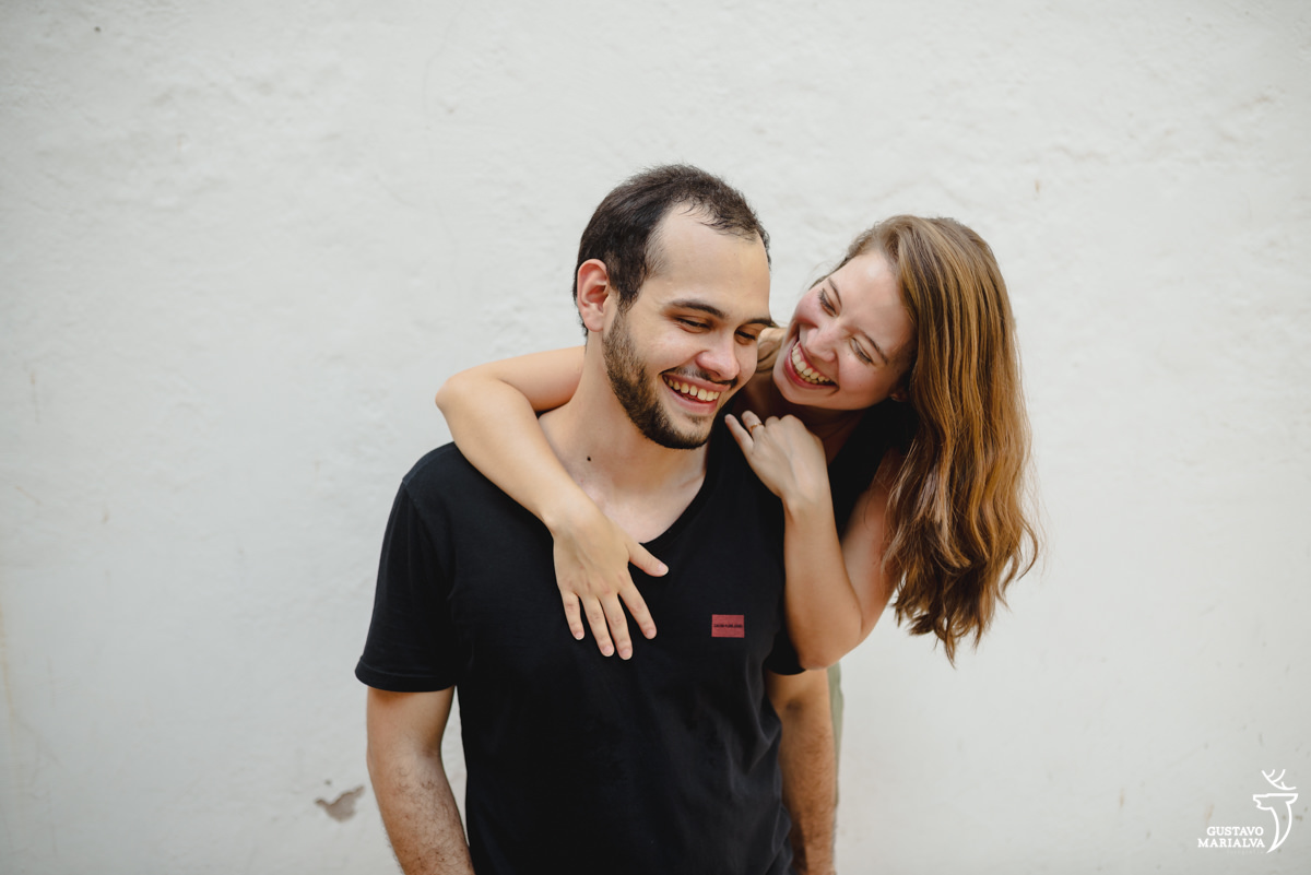 noiva abraça noivo que usa camisa preta e sorri em frente a parede branca