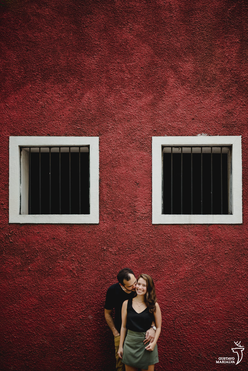 namorado usando camisa preta abraça namorada por trás em frente a parede vermelha