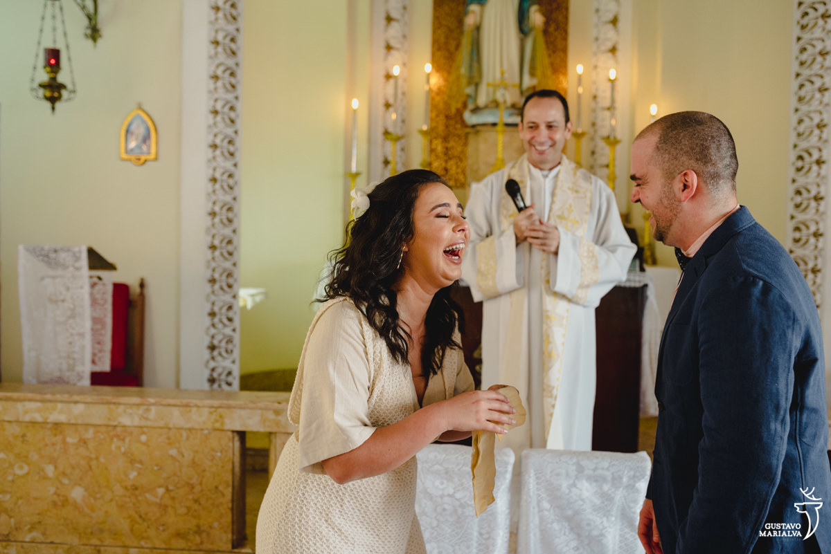 noiva sorri enquanto segura lenço bege para enxugar as lágrimas enquanto é observada pelo noivo que usa terno azul 