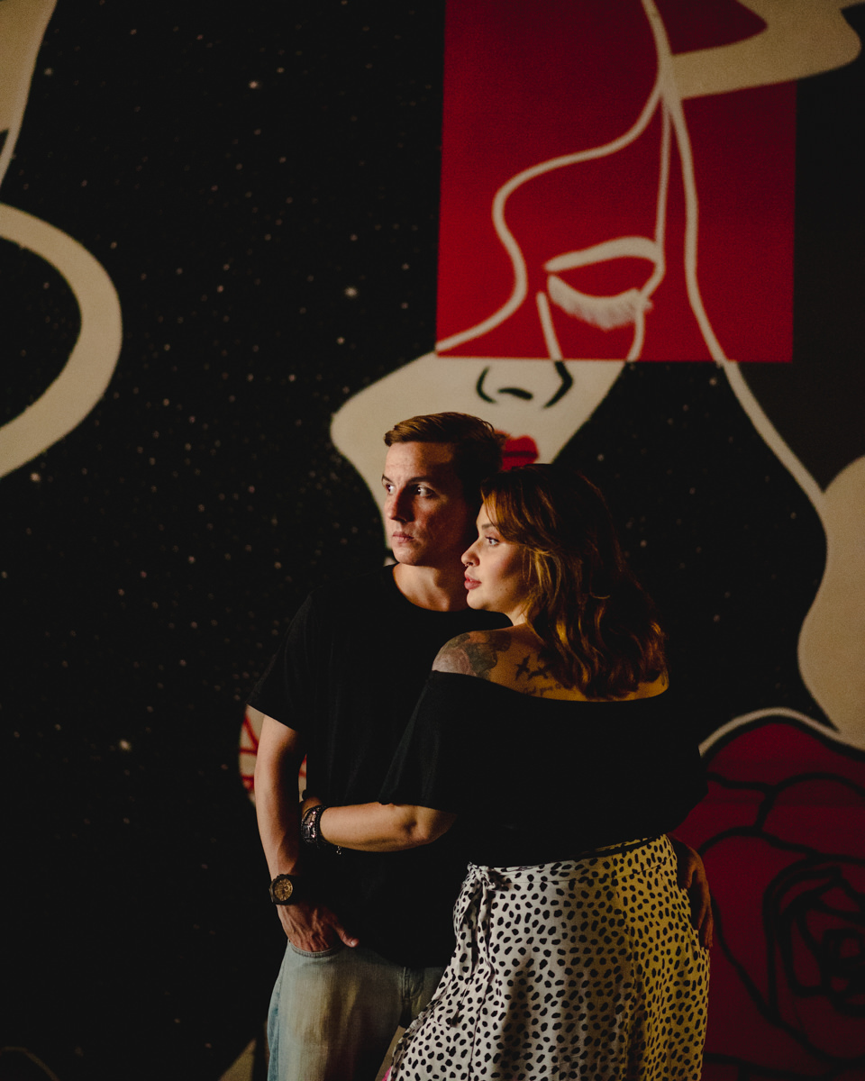 casal de camisa preta posa em frente a mural de uma mulher pintada de vermelho, branco e preto. 