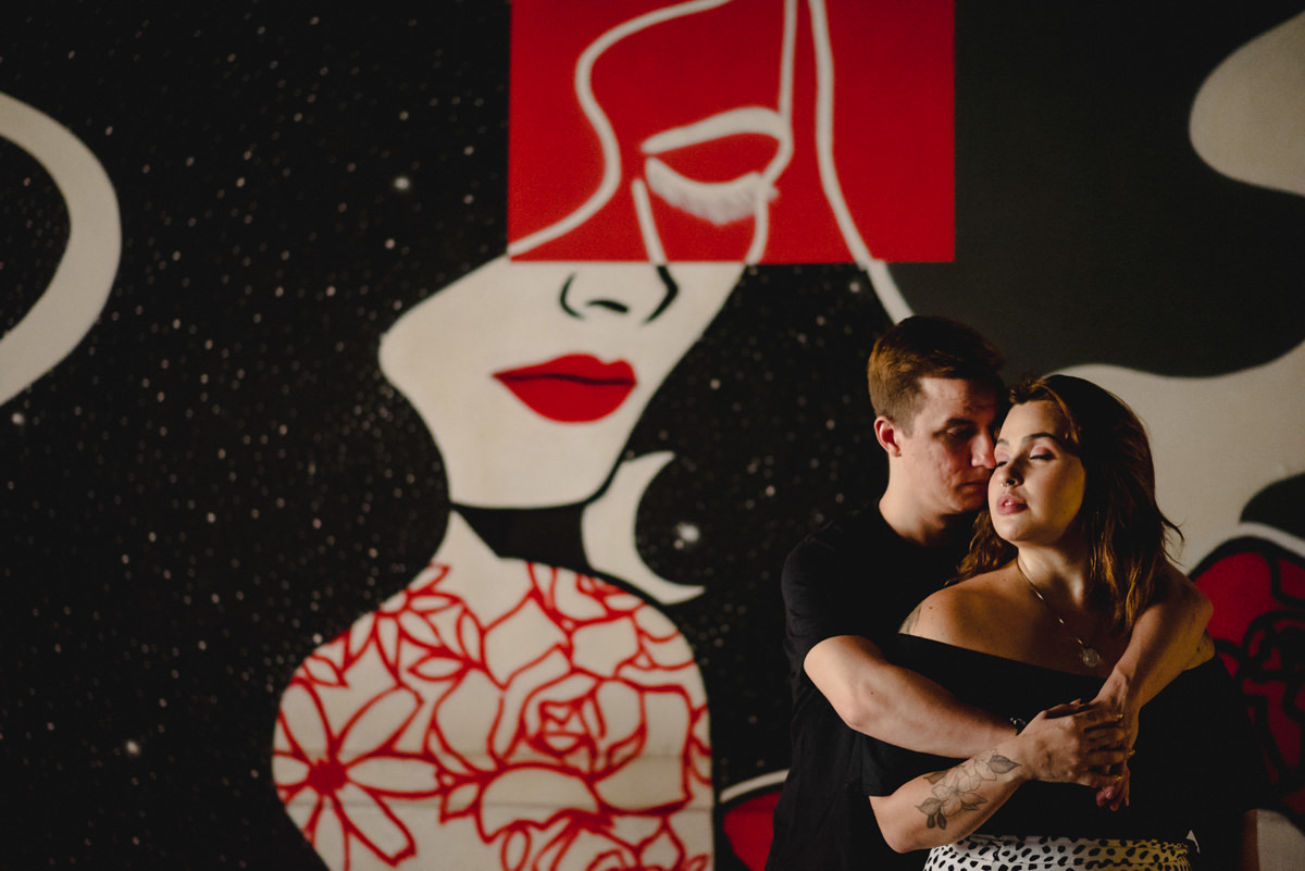 namorado de camisa preta abraça a namorada por trás, que também usa camisa preta, em frente a mural de uma mulher pintada de vermelho, branco e preto. 