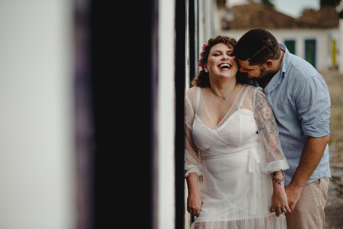 noivo de camisa azul e bermuda bege beija pescoço da noiva que usa vestido branco e sorri