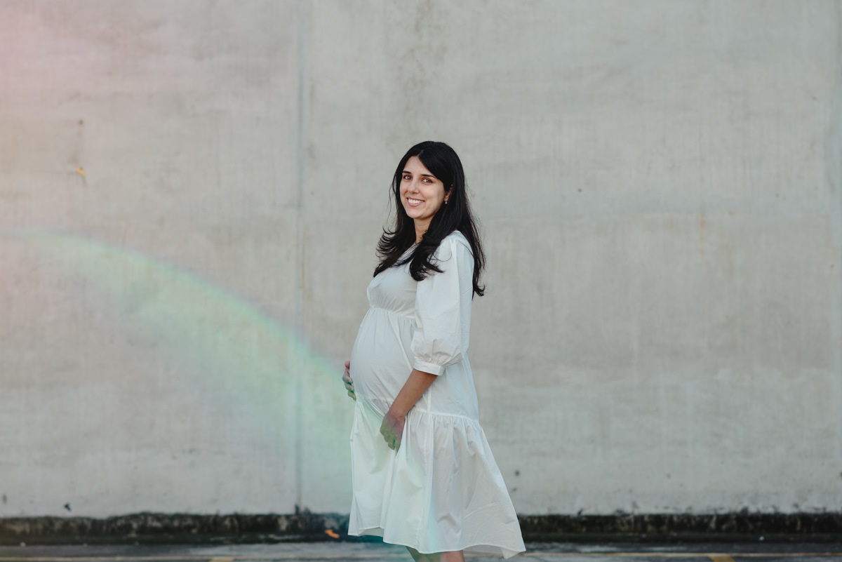 gestante de vestido branco posa em frente a parede branca com flaire iluminando