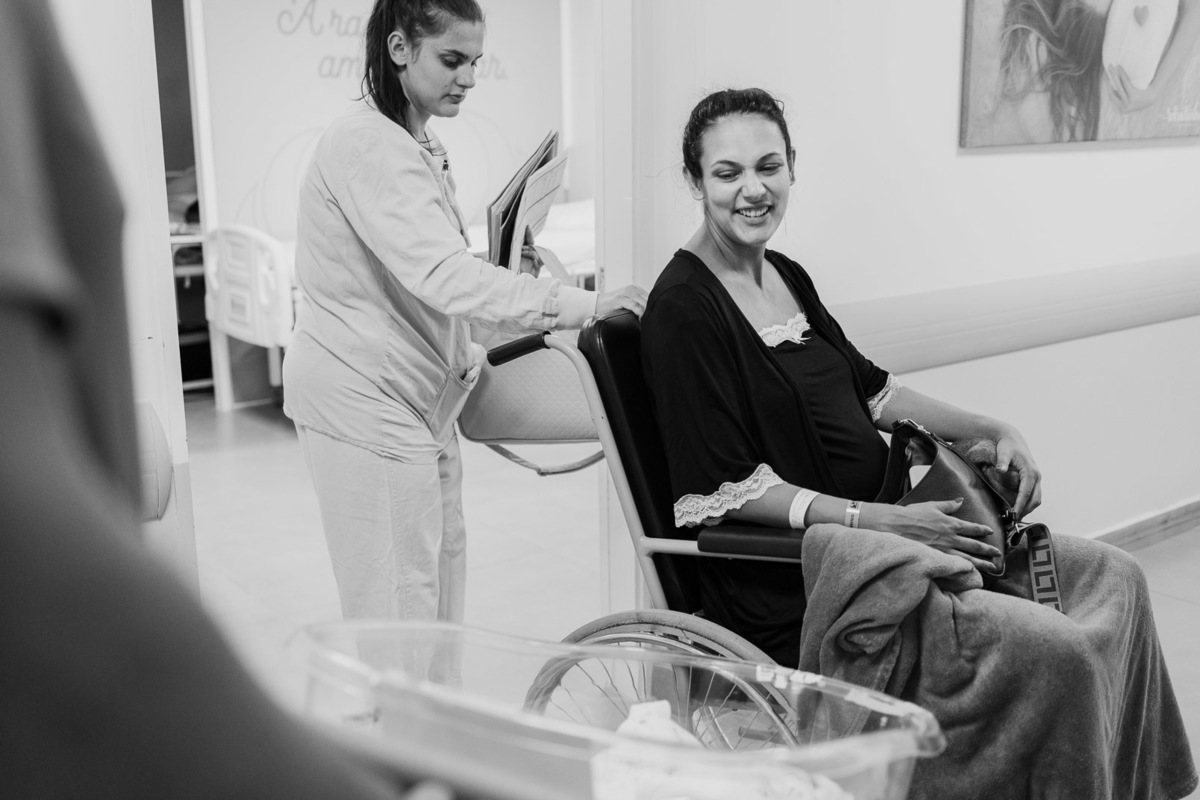 Mãe sorrindo em cadeira de rodas após parto no Hospital Bruno Born em Lajeado, acompanhada por enfermeira – foto de nascimento por Alba Estúdio