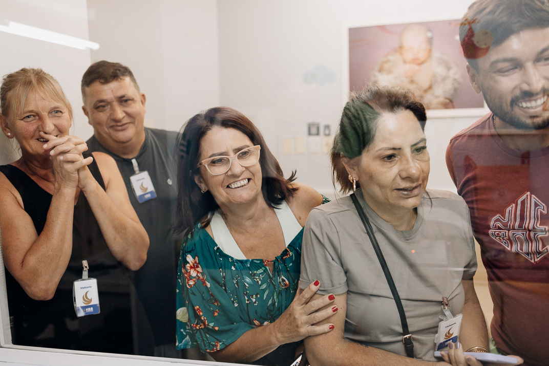 “Parentes e amigos registram com celulares a chegada do recém‑nascido através da divisória de vidro na maternidade”